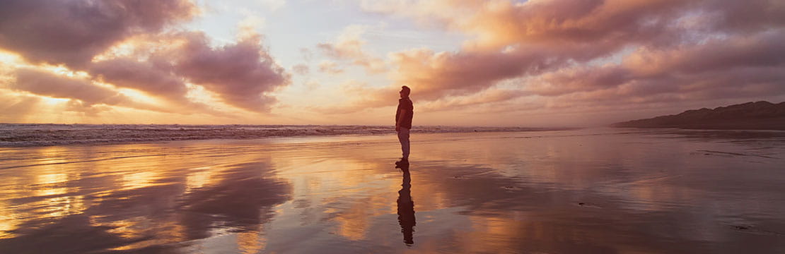 Person standing on a reflective beach at sunset, with colorful clouds and sky mirrored in the water.